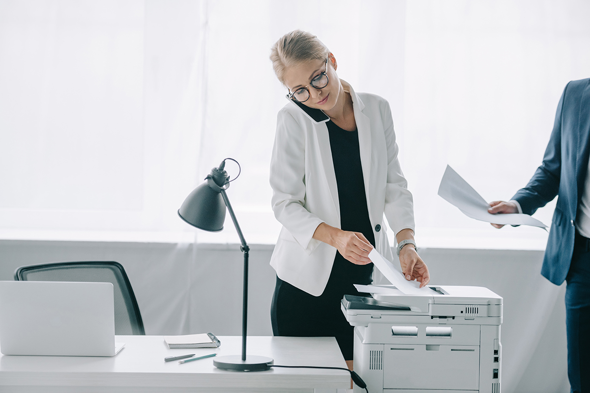 businesswoman talking on smartphone while using printer in office with colleague near by